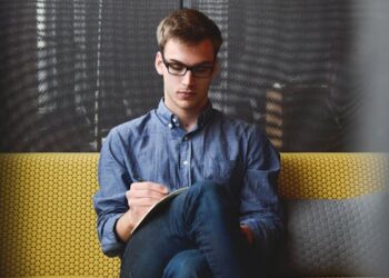 A young man in glasses writes in a notebook while sitting on a stylish couch indoors.