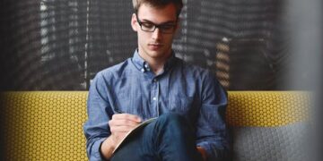 A young man in glasses writes in a notebook while sitting on a stylish couch indoors.