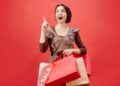 Cheerful woman with shopping bags celebrating a sale on a vibrant red backdrop.
