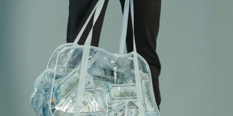 Individual in black attire holds a clear bag of cash, standing on scattered money indoors.