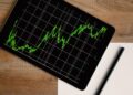 Flatlay of an iPad displaying stock market graph on a wooden desk with a pencil and paper.