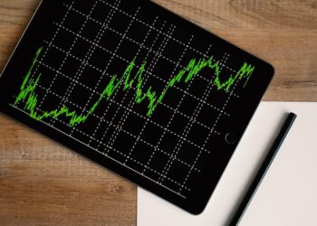 Flatlay of an iPad displaying stock market graph on a wooden desk with a pencil and paper.